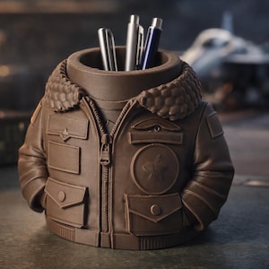 May include: A brown pen holder in the shape of a bomber jacket with a faux fur collar. The pen holder has a zip and pockets, and holds several pens. The background is blurred, suggesting an outdoor setting.