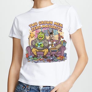 May include: White t-shirt with a cartoon illustration of Shrek and Donkey on a purple sofa, with the text "TWO GROWN MEN, ZERO MATURITY." The characters are gaming and eating pizza. The design includes a lamp and scattered items.