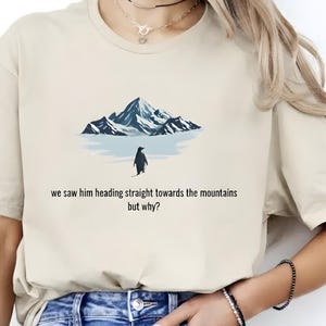 Könnte beinhalten: Beiges T-Shirt mit einem Pinguin, der auf eine Bergkette zugeht. Unter dem Bild steht der Text "we saw him heading straight towards the mountains but why?". Ein lässiges Kleidungsstück für den Alltag.