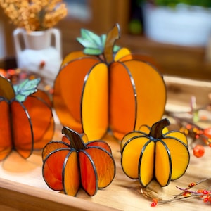 May include: A collection of three-dimensional stained glass pumpkins in varying shades of orange and yellow, each featuring dark soldered seams and small green glass leaves attached to the top stems, arranged on a wooden surface.