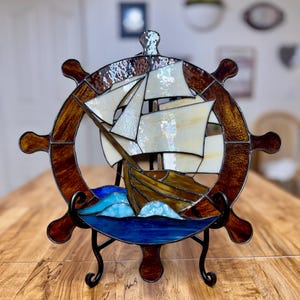 Könnte beinhalten: Ein Buntglas-Suncatcher in Form eines Schiffssteuerrads mit einem braunen, holztexturierten Rahmen, einem zentralen Segelschiff mit cremeweißen Segeln und einer Basis aus tiefblauen und helltürkisfarbenen Glaswellen auf einem schwarzen Metallständer.