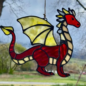Op de afbeelding: Gebrandschilderde glazen draak zonnevanger met een rode body, gele vleugels en een rode kop. De draak hangt aan een bruin touwtje. De vleugels van de draak zijn lichtgeel met zwarte contouren. De draak is een decoratief item.