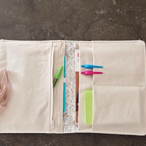 Può includere: Copertina per notebook in tela color crema, aperta per rivelare un interno a fantasia floreale, una tasca con cerniera e diverse altre tasche. All'interno ci sono una penna verde, una penna blu, una penna rosa, un taccuino turchese e un blocco note verde. Un nastro è attaccato.