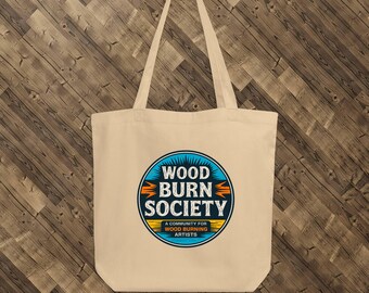 Könnte beinhalten: Beige Tragetasche mit dem Schriftzug "WOOD BURN SOCIETY" in einem blauen, orangefarbenen und gelben kreisförmigen Design. Der Text "A COMMUNITY FOR WOOD BURNING ARTISTS" befindet sich darunter. Die Tasche hat lange Henkel und hängt an einem schwarzen Haken.