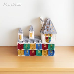 May include: A crocheted advent calendar in the shape of a castle, featuring a grey roof and white walls. It has 12 numbered pockets in colours like red, green, and blue. A crest is visible on the front of the castle.