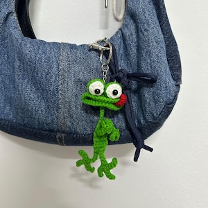 Könnte beinhalten: Ein grüner gehäkelter Frosch-Schlüsselanhänger mit weißen Augen und einem roten Mund hängt an einem silbernen Verschluss, der an einer Jeans-Tasche befestigt ist. Der Frosch hat ausgestreckte Arme und Beine. Die Tasche ist aus blauem Jeansstoff.