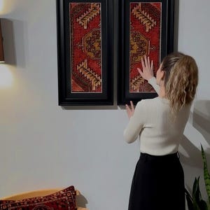 May include: Two framed rectangular wall art pieces with a red and gold patterned design. The frames are black. A matching pillow is on a chair. The overall theme is home decor.