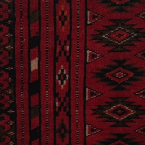 Puede incluir: Una pieza textil enmarcada con un fondo rojo intenso y bandas verticales de patrones geom&eacute;tricos en negro y marr&oacute;n oscuro, incluyendo rombos y tri&aacute;ngulos. La obra est&aacute; rodeada por un paspart&uacute; negro y un marco de madera de color blanco.