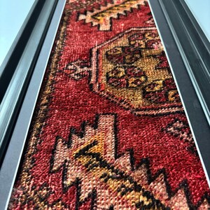Pode incluir: Tapete vintage emoldurado com um padr&atilde;o geom&eacute;trico vermelho, amarelo e preto. O tapete &eacute; exibido em uma moldura preta, criando um contraste marcante. A textura e as cores do tapete s&atilde;o bem definidas, mostrando seu design intrincado.