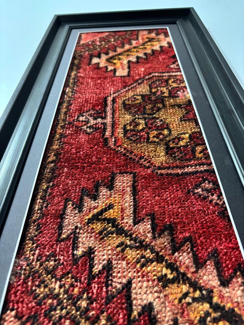 Pode incluir: Tapete vintage emoldurado com um padr&atilde;o geom&eacute;trico vermelho, amarelo e preto. O tapete &eacute; exibido em uma moldura preta, criando um contraste marcante. A textura e as cores do tapete s&atilde;o bem definidas, mostrando seu design intrincado.