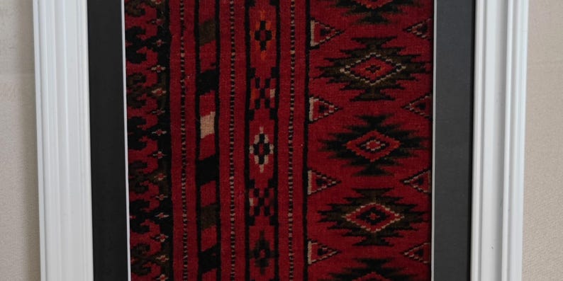 Puede incluir: Una pieza textil enmarcada con un fondo rojo intenso y complejos patrones geom&eacute;tricos en negro y blanco. La tela se exhibe dentro de un paspart&uacute; negro y un marco de madera blanca, mostrando rayas verticales y motivos en forma de diamante.