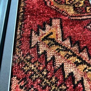 Pode incluir: Close-up de um tapete antigo emoldurado com um padr&atilde;o geom&eacute;trico detalhado. O tapete apresenta uma rica paleta de cores vermelha, preta e dourada. O design intrincado &eacute; emoldurado por uma borda cinza escuro, destacando a textura e o artesanato do tapete.
