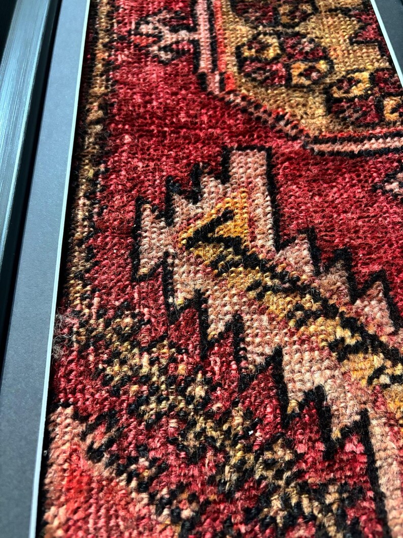 Pode incluir: Close-up de um tapete antigo emoldurado com um padr&atilde;o geom&eacute;trico detalhado. O tapete apresenta uma rica paleta de cores vermelha, preta e dourada. O design intrincado &eacute; emoldurado por uma borda cinza escuro, destacando a textura e o artesanato do tapete.