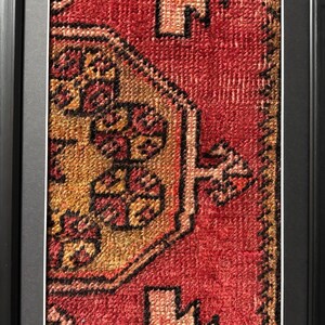 Pode incluir: Close-up de um tapete retangular emoldurado com um design tradicional. O tapete apresenta um fundo vermelho com padr&otilde;es intrincados em tons de ouro, castanho e preto. A moldura &eacute; preta e tem um design simples e moderno.
