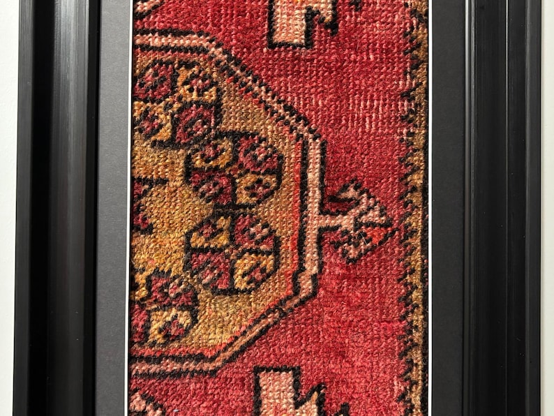 Pode incluir: Close-up de um tapete retangular emoldurado com um design tradicional. O tapete apresenta um fundo vermelho com padr&otilde;es intrincados em tons de ouro, castanho e preto. A moldura &eacute; preta e tem um design simples e moderno.
