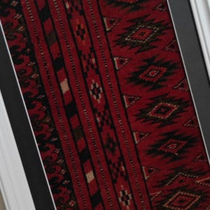 Puede incluir: Una pieza textil enmarcada con un fondo rojo intenso y complejos patrones geom&eacute;tricos en negro y crema. La tela rectangular est&aacute; montada sobre un paspart&uacute; gris oscuro y encerrada en un marco blanco sencillo, mostrando motivos repetitivos.
