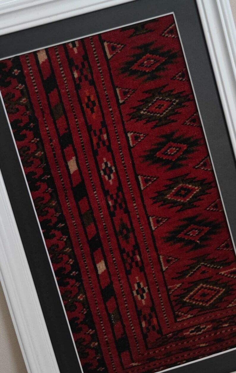 Puede incluir: Una pieza textil enmarcada con un fondo rojo intenso y complejos patrones geom&eacute;tricos en negro y crema. La tela rectangular est&aacute; montada sobre un paspart&uacute; gris oscuro y encerrada en un marco blanco sencillo, mostrando motivos repetitivos.