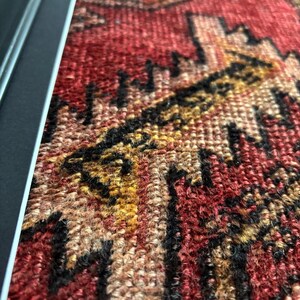 Pode incluir: Close-up de um tapete emoldurado em estilo vintage com um padr&atilde;o tecido detalhado. O tapete apresenta uma rica paleta de cores de vermelho, preto, dourado e bege. O design intrincado &eacute; emoldurado em uma moldura preta e prateada, mostrando a textura e o artesanato do tapete.