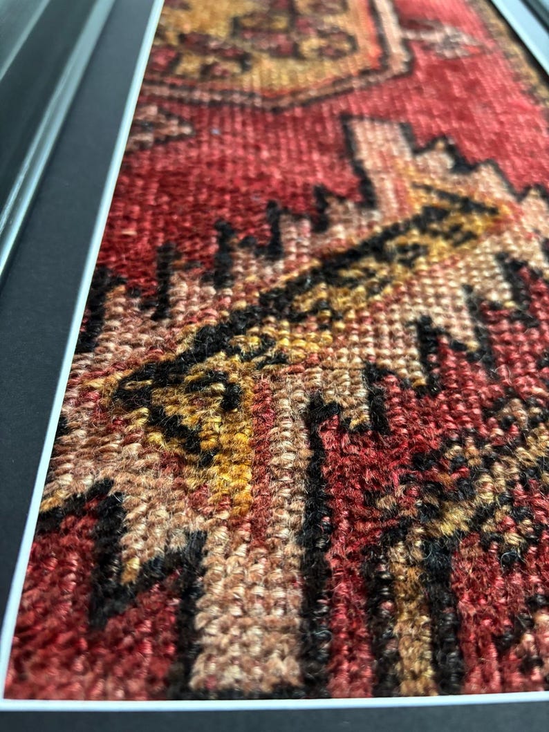 Pode incluir: Close-up de um tapete emoldurado em estilo vintage com um padr&atilde;o tecido detalhado. O tapete apresenta uma rica paleta de cores de vermelho, preto, dourado e bege. O design intrincado &eacute; emoldurado em uma moldura preta e prateada, mostrando a textura e o artesanato do tapete.
