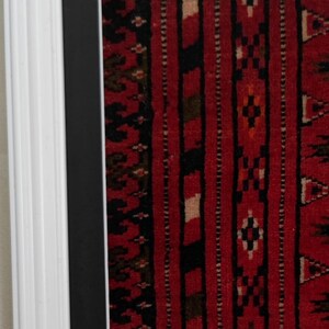 Puede incluir: Una pieza textil enmarcada que presenta un fondo rojo vibrante con rayas verticales en negro, blanco y verde oscuro. La tela muestra intrincados patrones geom&eacute;tricos en toda su superficie. La pieza est&aacute; dentro de un marco de madera blanco.