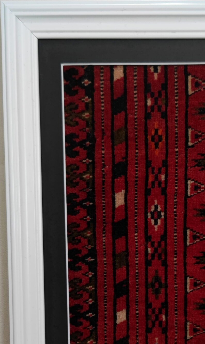 Puede incluir: Una pieza textil enmarcada que presenta un fondo rojo vibrante con rayas verticales en negro, blanco y verde oscuro. La tela muestra intrincados patrones geom&eacute;tricos en toda su superficie. La pieza est&aacute; dentro de un marco de madera blanco.