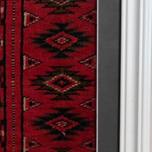 Puede incluir: Una pieza textil enmarcada con un fondo rojo intenso y patrones geom&eacute;tricos de diamantes en negro y marr&oacute;n oscuro. El textil est&aacute; rodeado por un paspart&uacute; gris oscuro y un marco de madera blanca brillante con un dise&ntilde;o de perfil escalonado.