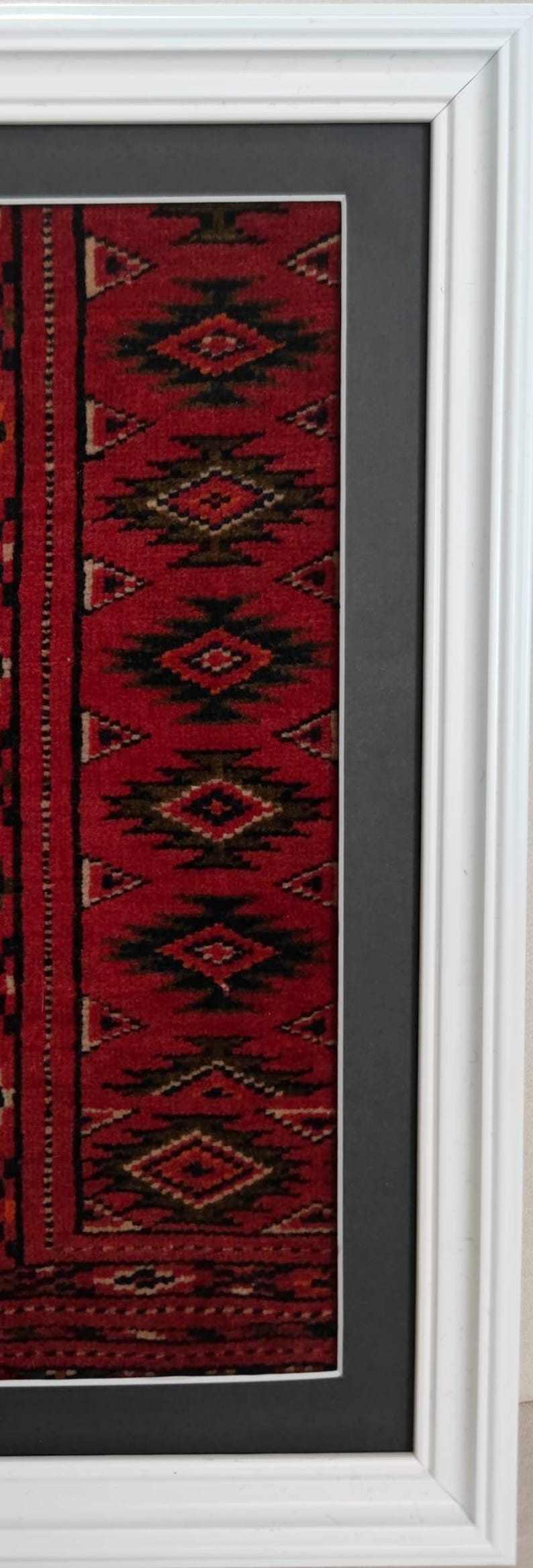 Puede incluir: Una pieza textil enmarcada con un fondo rojo intenso y patrones geom&eacute;tricos de diamantes en negro y marr&oacute;n oscuro. El textil est&aacute; rodeado por un paspart&uacute; gris oscuro y un marco de madera blanca brillante con un dise&ntilde;o de perfil escalonado.