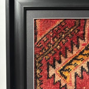 Pode incluir: Sec&ccedil;&atilde;o emoldurada de um tapete com padr&atilde;o vermelho, laranja e preto. O design do tapete apresenta formas geom&eacute;tricas e uma apar&ecirc;ncia texturizada. A moldura &eacute; preta e tem um design simples e moderno, com uma moldura branca ao redor do tapete.