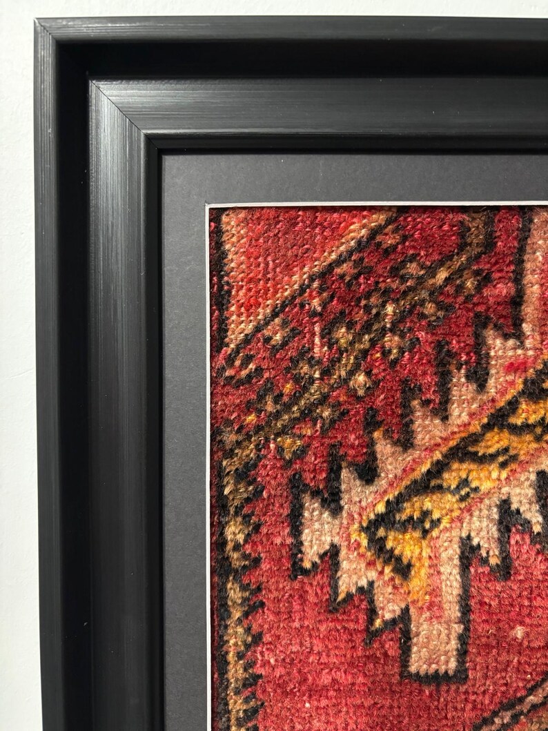 Pode incluir: Sec&ccedil;&atilde;o emoldurada de um tapete com padr&atilde;o vermelho, laranja e preto. O design do tapete apresenta formas geom&eacute;tricas e uma apar&ecirc;ncia texturizada. A moldura &eacute; preta e tem um design simples e moderno, com uma moldura branca ao redor do tapete.