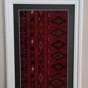 Puede incluir: Una pieza de arte mural rectangular que presenta un fragmento de tejido rojo, negro y blanco con patrones geom&eacute;tricos de diamantes, encerrado en un paspart&uacute; negro y un marco de madera blanca. El tejido muestra rayas verticales y motivos.