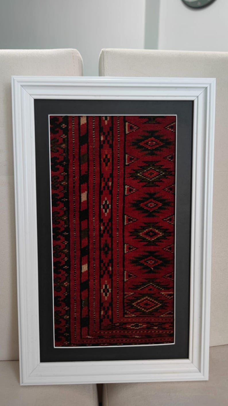 Puede incluir: Una pieza de arte mural rectangular que presenta un fragmento de tejido rojo, negro y blanco con patrones geom&eacute;tricos de diamantes, encerrado en un paspart&uacute; negro y un marco de madera blanca. El tejido muestra rayas verticales y motivos.