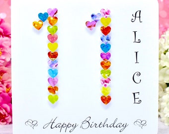 Può includere: Un biglietto d'auguri bianco con il numero 11 composto da piccoli ornamenti colorati a forma di cuore. Il nome ALICE è stampato verticalmente in nero sul lato destro, con la scritta Happy Birthday nella parte inferiore.