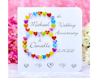 Può includere: Un biglietto bianco con un design a forma di cuore colorato che compone il numero otto. Il biglietto dice "Michael & Danielle 8° Anniversario di Matrimonio 12.09.2021".