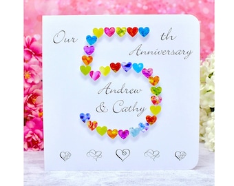 Può includere: Un biglietto bianco con un design a forma di cuore colorato che forma il numero 5. Il testo "Our 5th Anniversary Andrew & Cathy" è stampato sul biglietto.