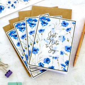 May include: Greeting cards with blue floral designs and the text "Just a Note to Say...". The cards are surrounded by brown envelopes, a gold pen, and binder clips. The cards feature a white background with blue flowers.