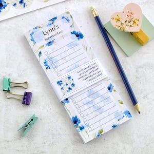 May include: A shopping list notepad featuring a blue floral pattern, with the text "Lynn's SHOPPING LIST". The notepad is accompanied by a pencil, a heart-shaped clip, and other small clips. The notepad has a band with text that reads "We hope you love your personalised notepad".