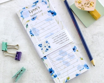 Può includere: Un blocco note per la lista della spesa personalizzato con un design floreale nei toni del blu. Il blocco note è tenuto insieme da una fascia abbinata e include una matita, una clip a forma di cuore e altre piccole clip. Il testo sul blocco note recita "Lynn's SHOPPING LIST".