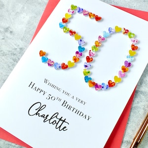 May include: A white card with the number 50 made out of colorful hearts. The text "Wishing you a very happy 50th birthday Charlotte" is printed on the card.