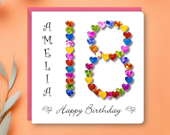 Può includere: Un biglietto di compleanno bianco con il nome "Amelia" in corsivo nero. Il numero "18" è formato da cuori colorati. Le parole "Happy Birthday" sono scritte sotto, con accenti a cuore.