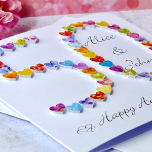 May include: A white card with a colourful heart design spelling out the number 50. The card says "Alice & John" and "Happy Anniversary".
