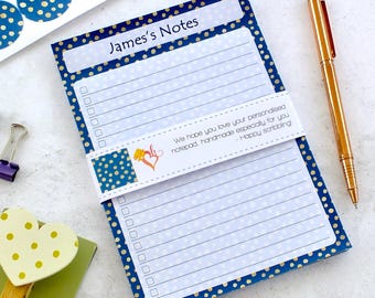 Può includere: Un blocco note personalizzato con il testo "James's Notes" in alto. Il blocco note ha un bordo a pois blu e una fascia abbinata con testo. Una penna dorata è appoggiata accanto al blocco note, insieme a adesivi abbinati.