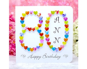 Può includere: Un biglietto bianco con il numero 80 fatto di cuori colorati. Il numero 80 è circondato dalle parole "Buon compleanno".
