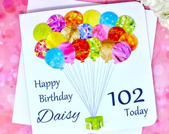 Può includere: Un biglietto bianco con un disegno di palloncini blu e bianchi. Il biglietto dice "Happy Birthday Daisy 102 Today".