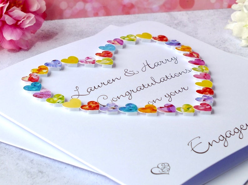 May include: A white card with a heart made of colorful paper hearts. The card says "Lauren & Harry Congratulations on your Engagement".