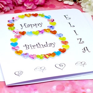 May include: A white card with the words "Happy Birthday" and a number 8 made of colorful hearts. The name "Eliza" is written vertically on the right side of the card.