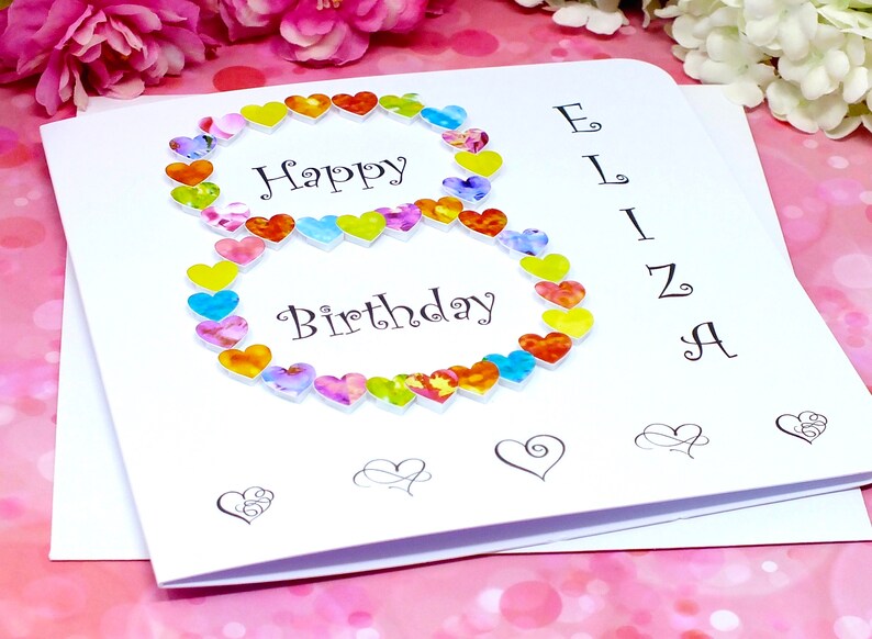 May include: A white card with the words "Happy Birthday" and a number 8 made of colorful hearts. The name "Eliza" is written vertically on the right side of the card.