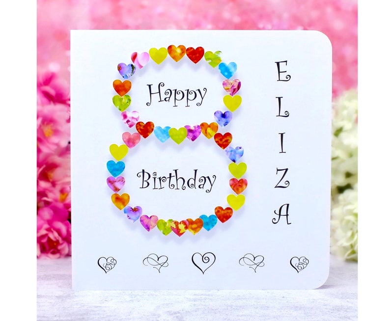 May include: A white card with a colorful heart-shaped number eight, the words "Happy Birthday" and the name "Eliza" spelled vertically. The card has five small hearts at the bottom.
