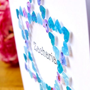 May include: A white card with a blue and purple heart border. The name Zacharia is written in black ink inside the heart border.