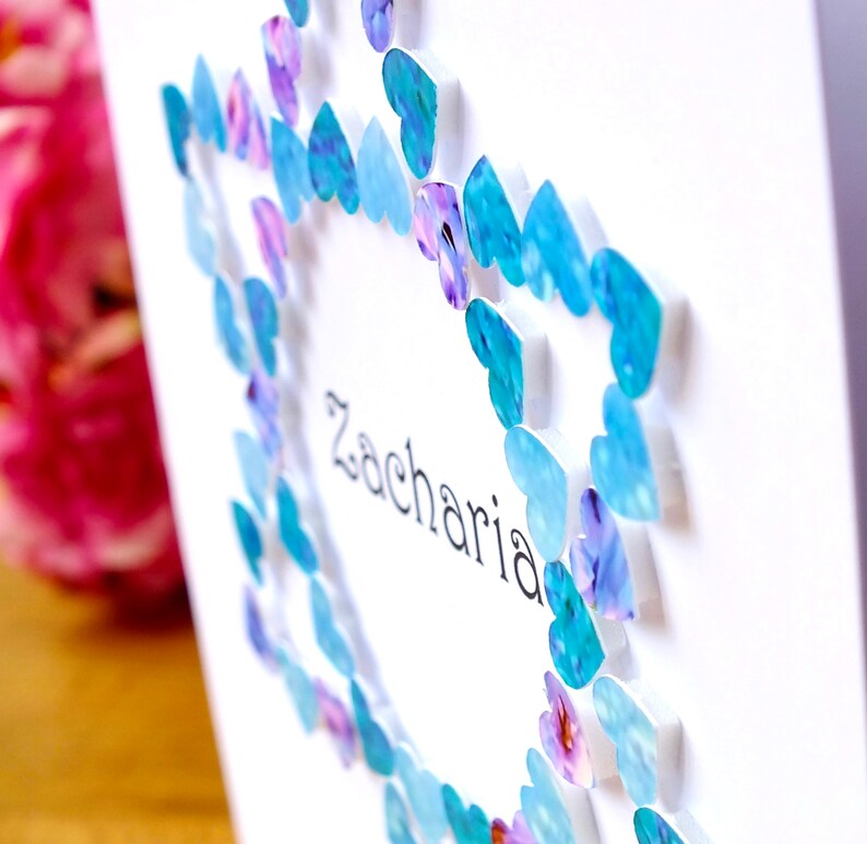 May include: A white card with a blue and purple heart border. The name Zacharia is written in black ink inside the heart border.
