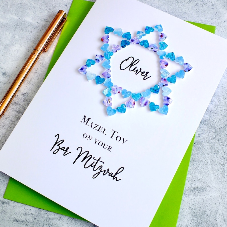May include: A white card with a blue and purple heart-shaped design in the shape of a star. The card says "Oliver" in the center and "Mazel Tov on your Bar Mitzvah" below the design.